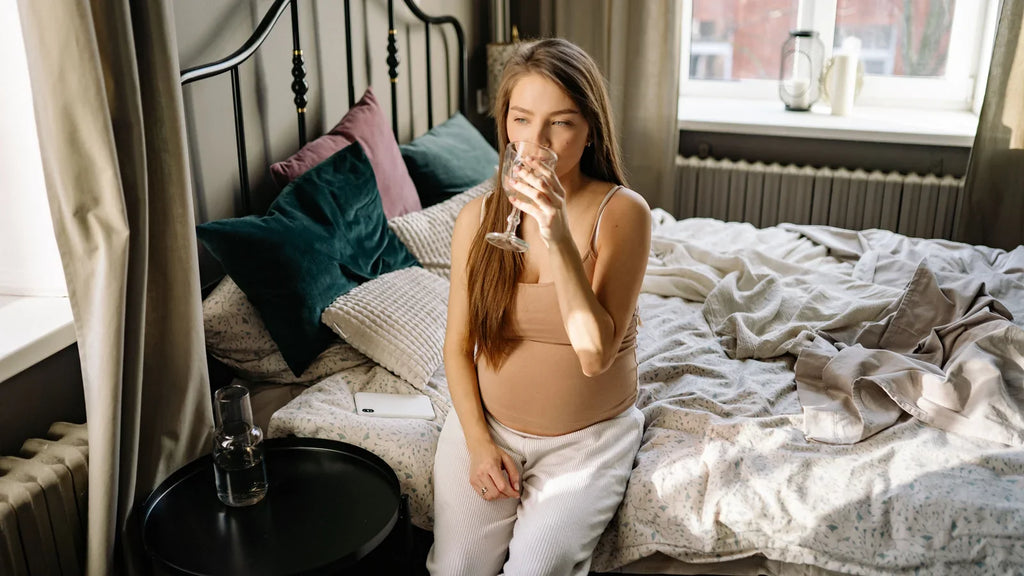 Femme enceinte assise sur un lit buvant un verre d’eau pendant une période de nausées